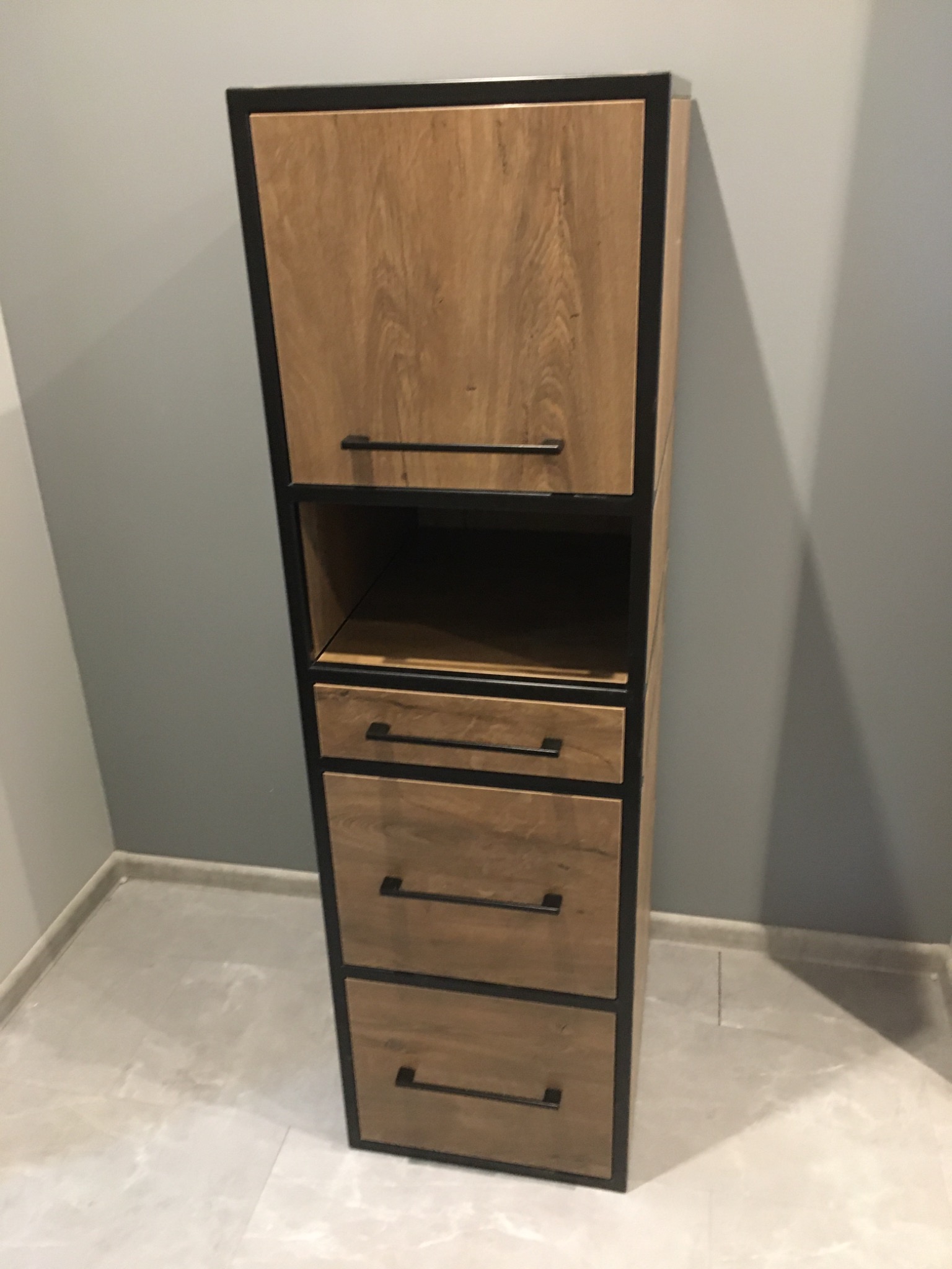 Wysoka, wąska szafka z drewnopodobnymi frontami i czarną metalową ramą, z trzema szufladami i otwartą półką, umieszczona w rogu pomieszczenia z szarą ścianą i jasną podłogą.