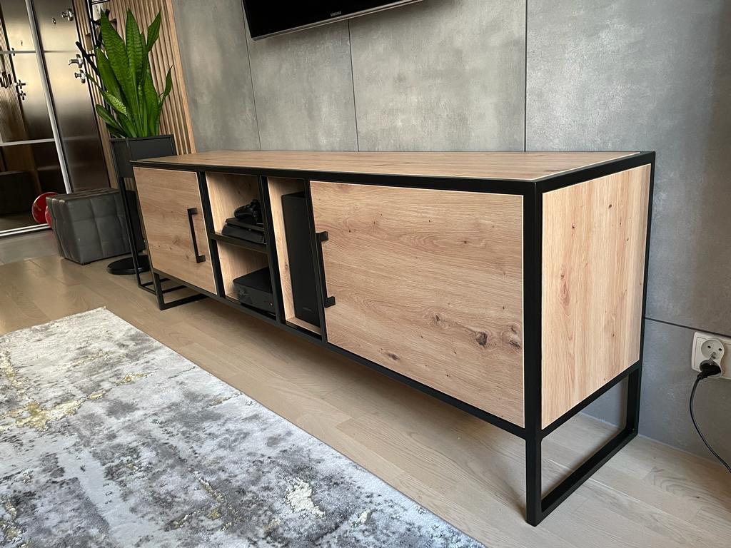 Nowoczesna szafka RTV z frontami z jasnego drewna i czarną metalową ramą, widoczne sprzęty RTV w otwartej półce, na tle szarej ściany i wzorzystego dywanu.