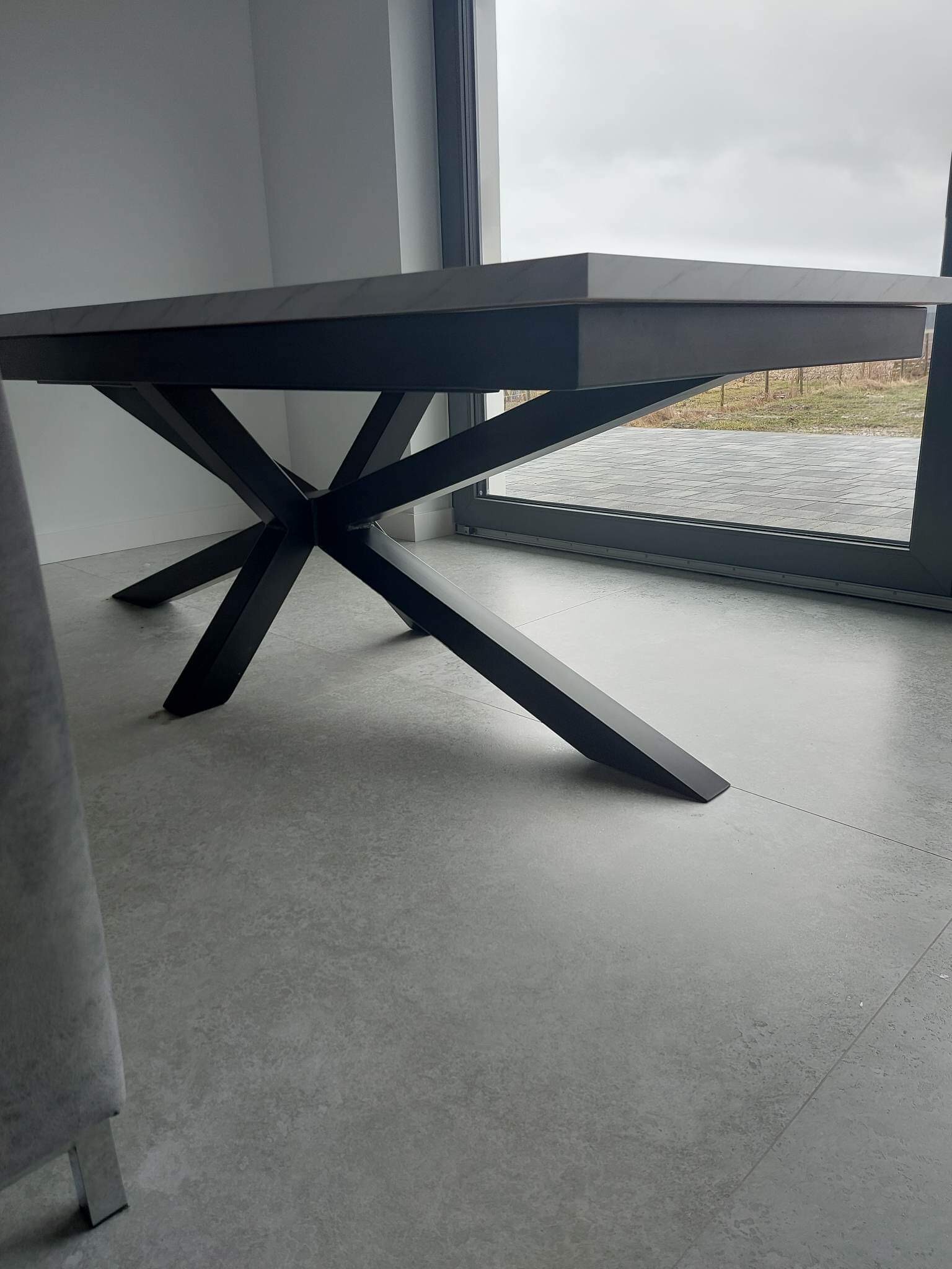 Stół z blatem imitującym marmur i geometryczną, czarną podstawą na tle okna tarasowego.