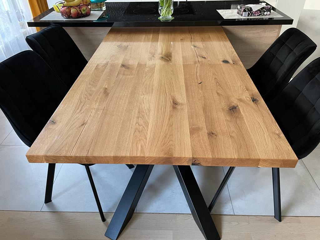 Rozkładany stół z jasnego drewna z czarnymi, metalowymi nogami w kształcie litery V, otoczony czarnymi krzesłami z pikowanym oparciem, na tle jasnej podłogi i blatu kuchennego z owocami.