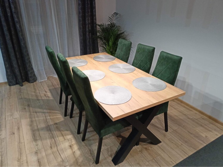 Stół z blatem w odcieniach drewna i sześcioma krzesłami z zieloną tapicerką, ustawiony na podłodze z paneli, z widoczną dekoracyjną rośliną w tle.