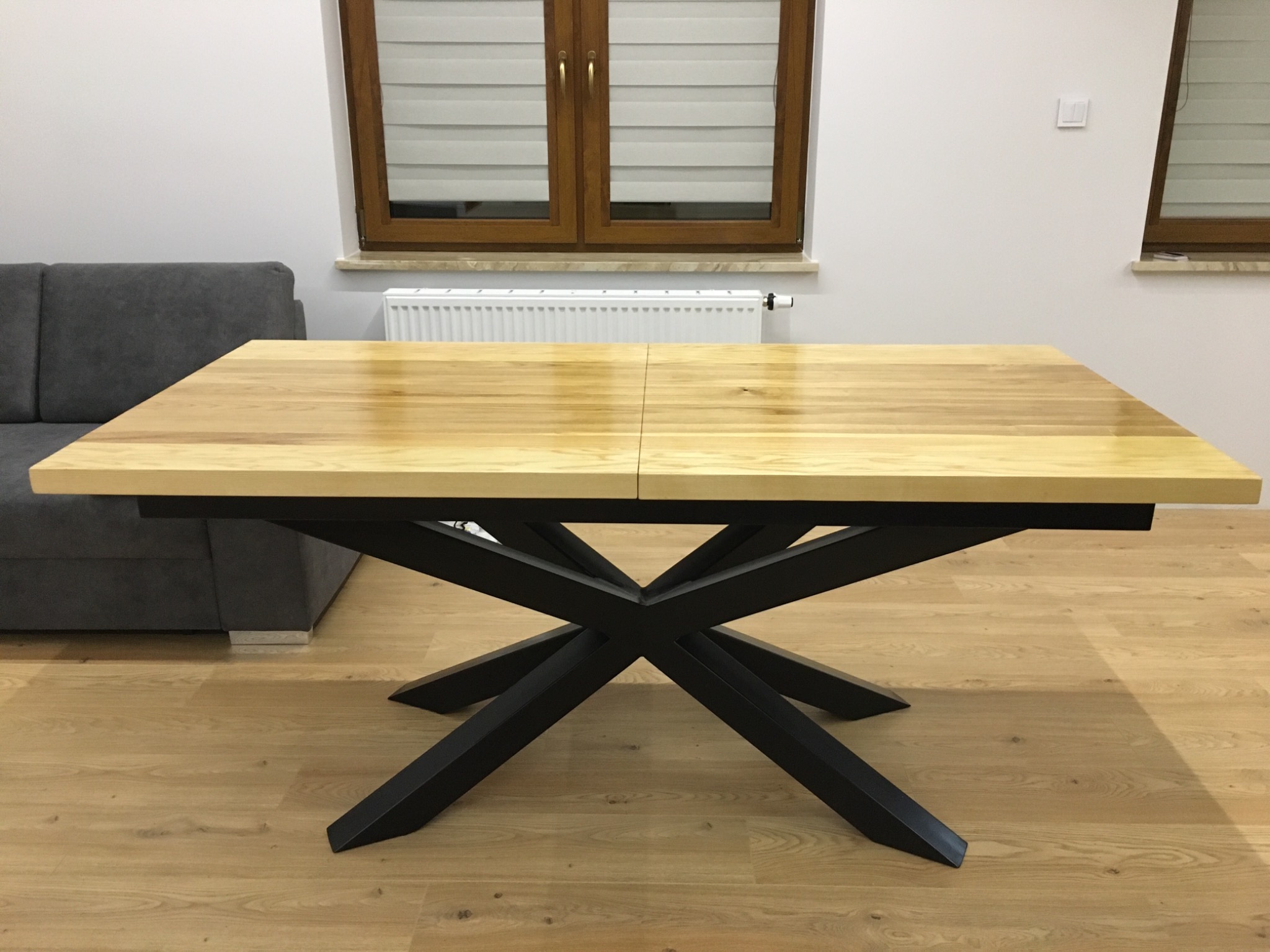 Rozkładany stół z jasnym drewnianym blatem i czarną, geometryczną podstawą w nowoczesnym salonie.
