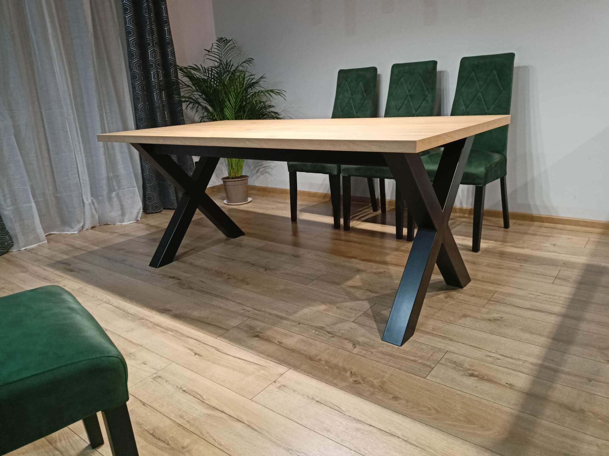 Stół z blatem z jasnego drewna na metalowych nogach w kształcie litery X, trzy krzesła tapicerowane zielonym welurem z przeszyciami w romby w tle, roślina doniczkowa i zasłony w geometryczny wzór.