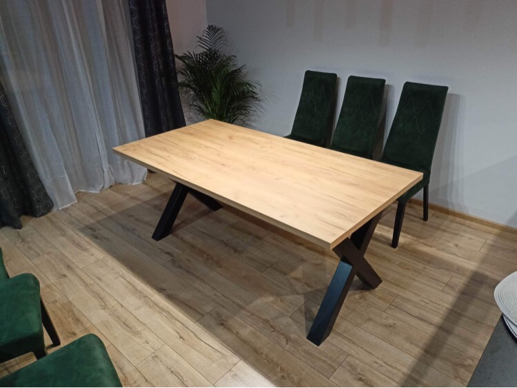 Stół jadalny z blatem w odcieniach drewna i czarnymi, krzyżowymi nogami, ustawiony w pomieszczeniu z drewnianą podłogą, zasłonami i rośliną doniczkową w tle, uzupełniony zielonymi krzesłami.