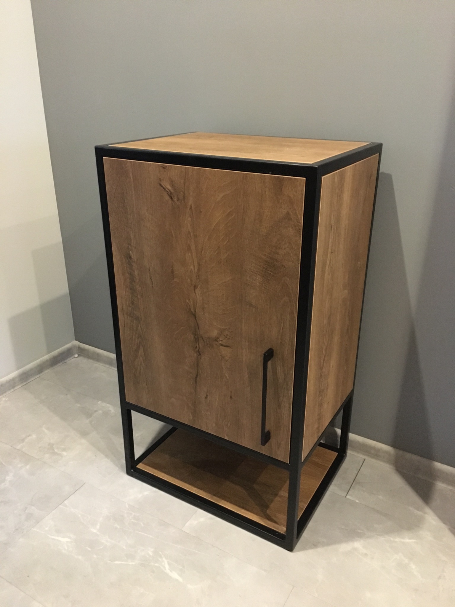 Szafka z drewnopodobną okleiną i czarną metalową ramą, z półką u dołu, na tle szarej ściany i marmurowej podłogi.