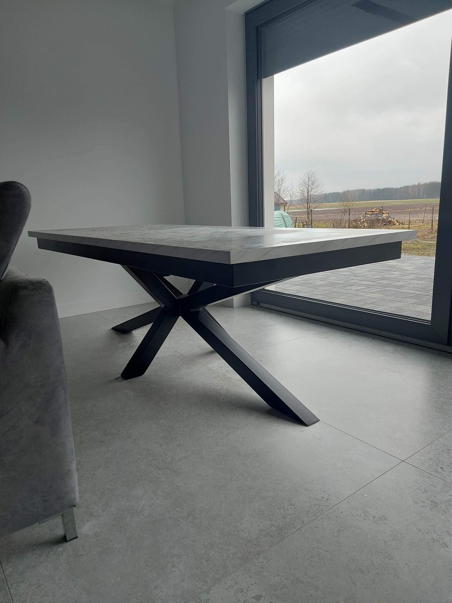 Rozkładany stół z blatem imitującym marmur i czarną, metalową podstawą w kształcie litery X, ustawiony na tle okna tarasowego z widokiem na stertę drewna.