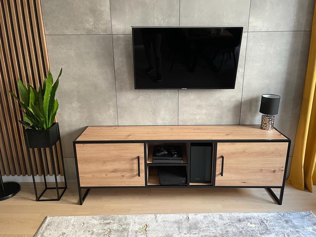 Nowoczesna szafka RTV z drewnianymi frontami i czarną metalową ramą, pod telewizorem na szarej ścianie, z rośliną w czarnej donicy po lewej stronie i zasłoną po prawej.