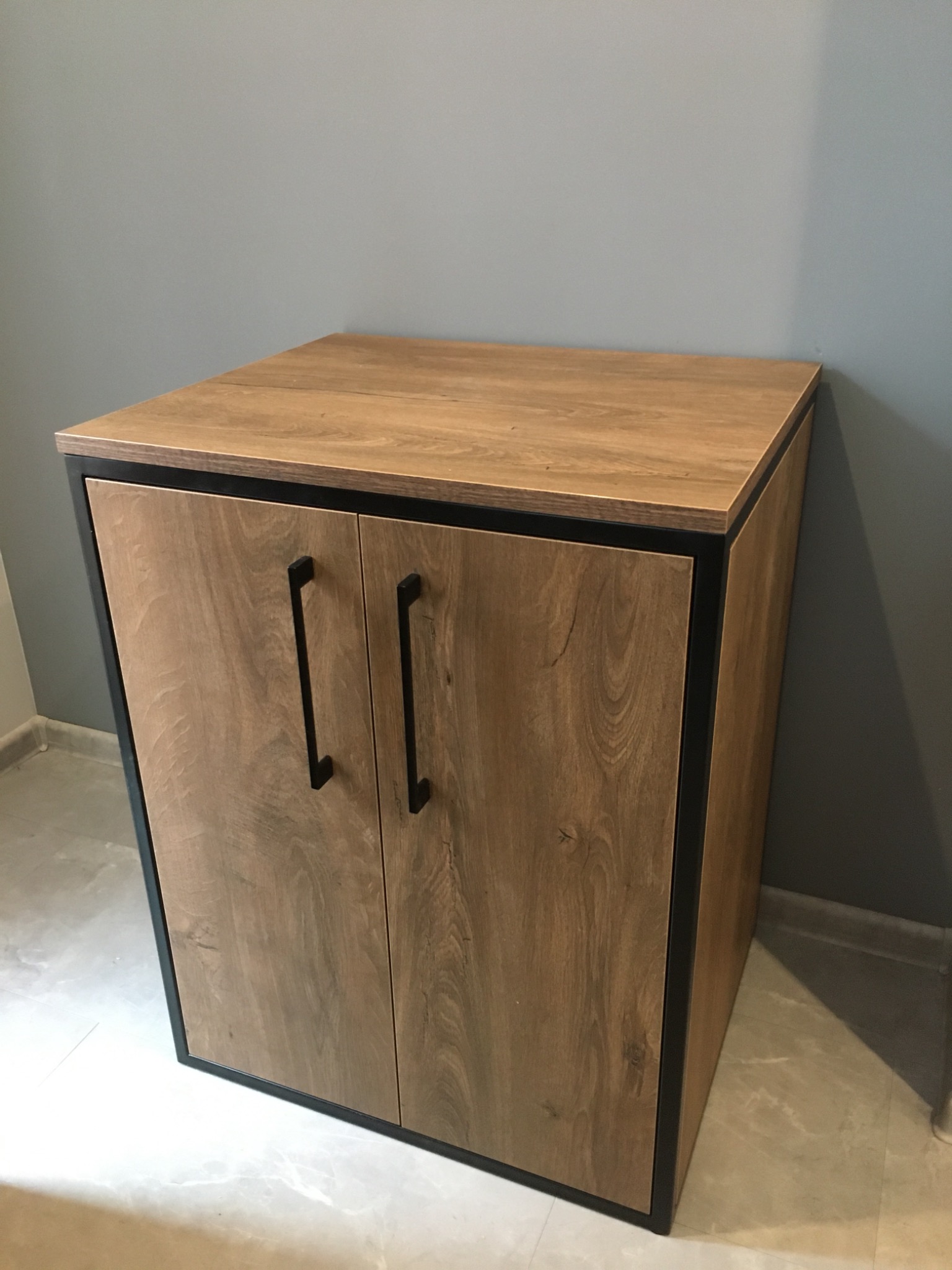 Mała, drewniana szafka z czarnymi, prostokątnymi uchwytami, otoczona czarną ramą, stojąca na marmurowej podłodze przy szarej ścianie.