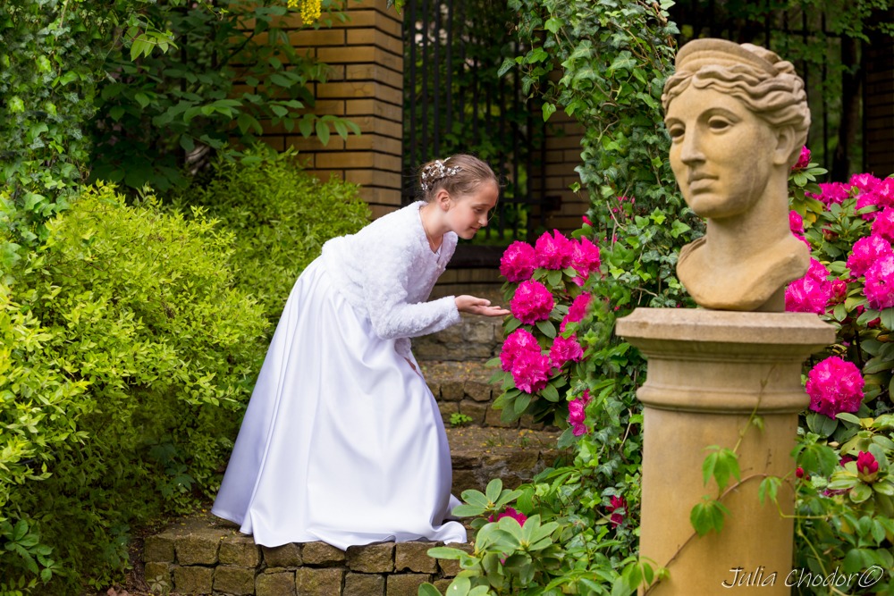 Dziewczynka w białej sukni, stojąca na kamiennych schodach w ogrodzie, wyciąga dłoń w stronę różowych kwiatów rododendronu, obok kamienny posąg głowy kobiety.