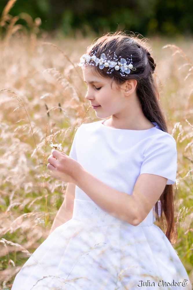 Portret dziewczynki w białej sukience komunijnej z wiankiem na głowie, trzymającej mały kwiatek na tle wysokich traw w złotym świetle.