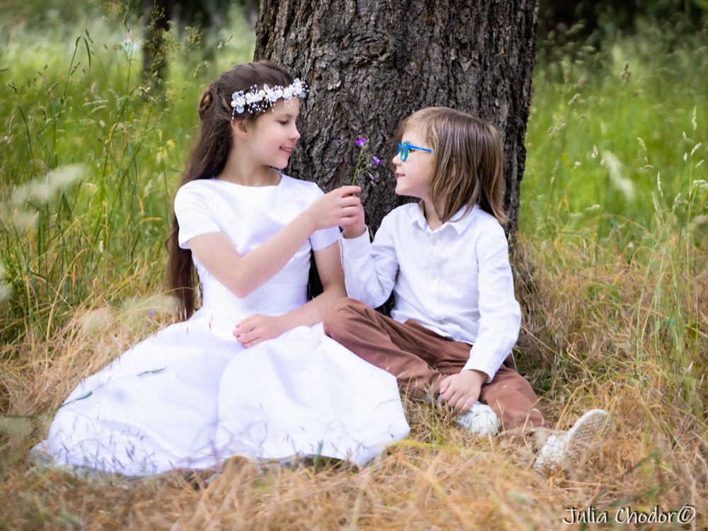 Dziewczynka w białej sukience i wianku oraz chłopiec w okularach siedzą pod drzewem, wymieniając się drobnym bukietem fioletowych kwiatów na tle łąki.