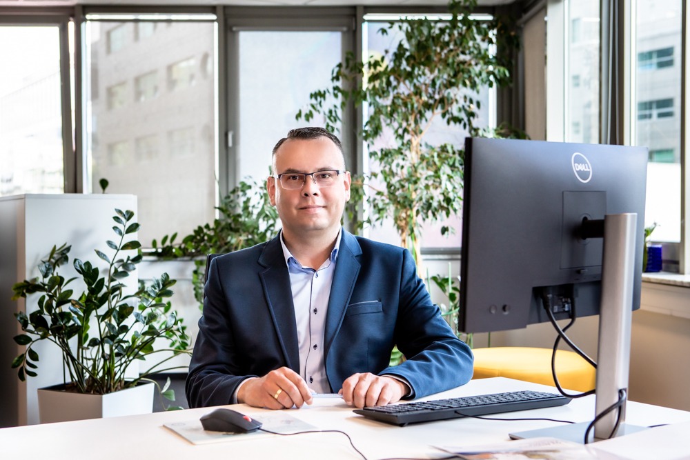 Portret mężczyzny w okularach i marynarce, siedzącego za biurkiem z komputerem w biurze w Warszawie, z roślinami doniczkowymi w tle.