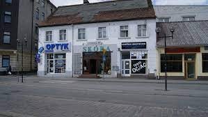 Fasada budynku z trzema lokalami usługowymi: Optyk z niebieskim szyldem i ekspozycją okularów, Skóra z wejściem i markizą, oraz lokal z napisem Tanie OC i reklamą ubezpieczeń komunikacyjnych.