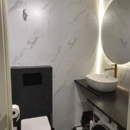 Nowoczesna łazienka z marmurowymi płytkami, owalnym lustrem z podświetleniem LED, umywalką nablatową z złotą armaturą, podwieszaną toaletą i pralką ukrytą pod blatem.