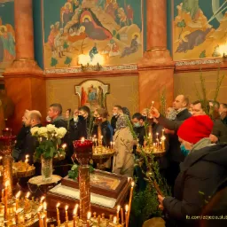 Wnętrze cerkwi podczas uroczystości religijnej: wierni trzymają zapalone świece i gałązki, w tle malowidła na ścianach i kolumnach.