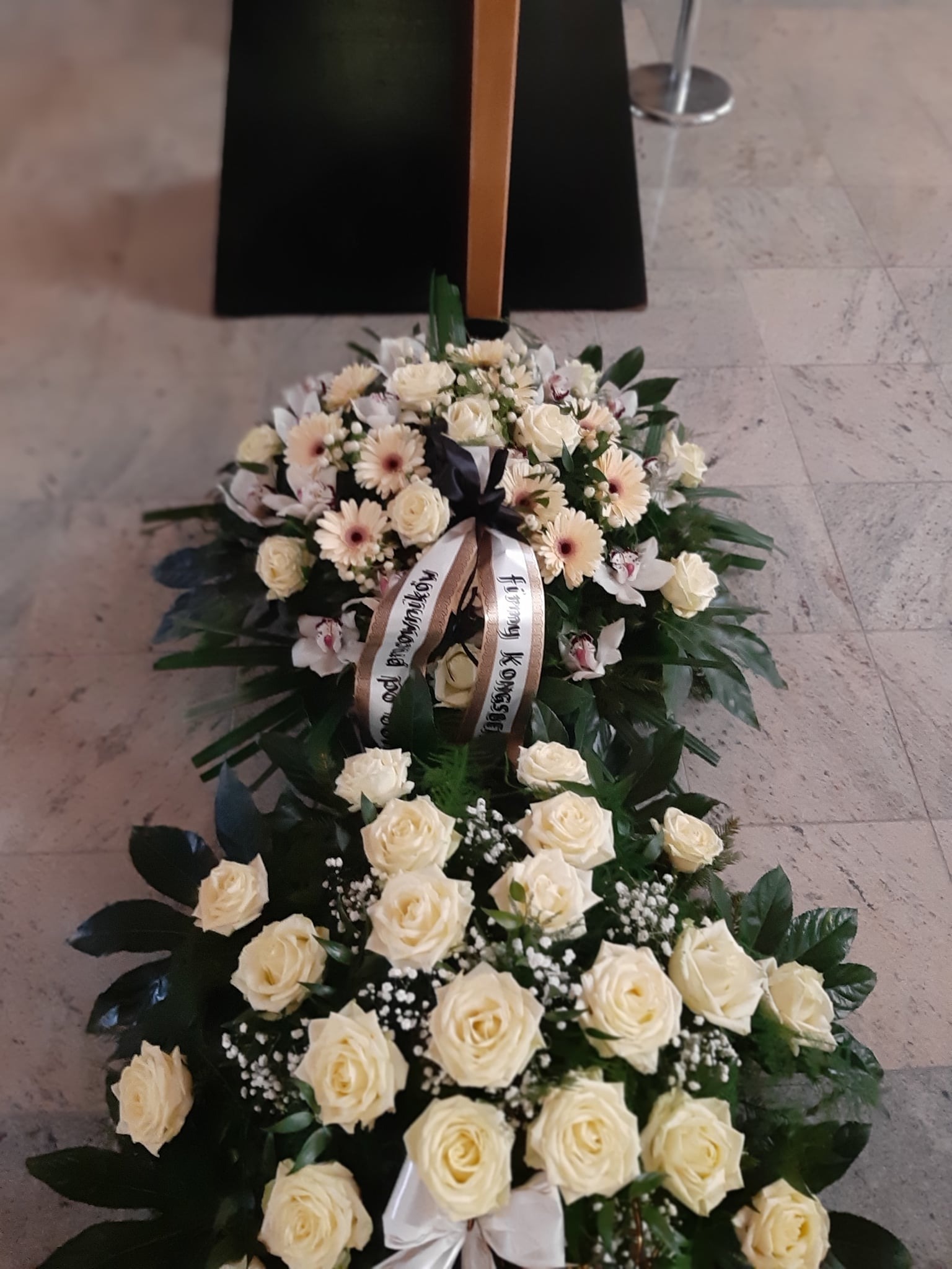 Kompozycja kwiatowa z białych róż, gerber i orchidei z wstęgą żałobną na marmurowej posadzce.