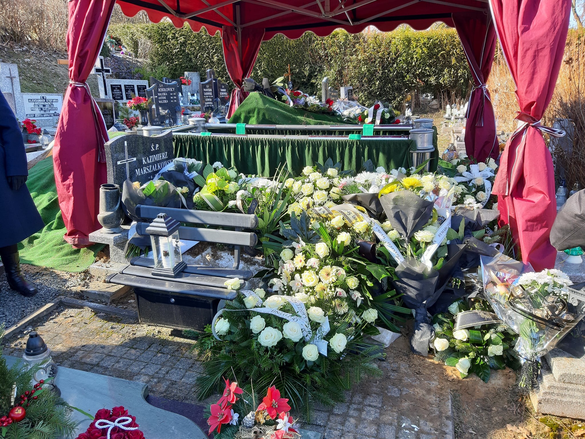 Grób przykryty zielonym materiałem pod czerwonym baldachimem, otoczony licznymi wiązankami białych kwiatów i zniczami na cmentarzu w słoneczny dzień.