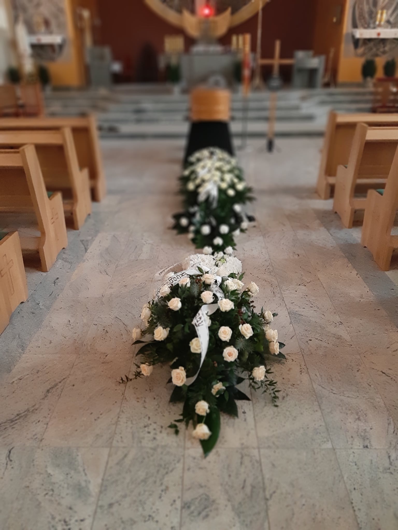 Dekoracja kwiatowa z białych róż i wstążek w kościele, ustawiona wzdłuż przejścia, prowadząca do trumny