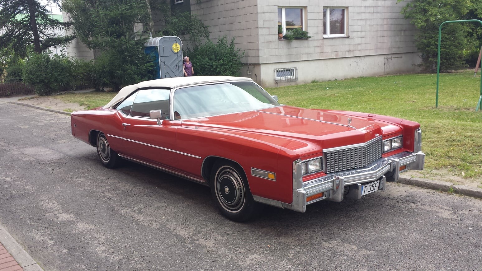 Czerwony kabriolet Cadillac Eldorado z białym dachem zaparkowany na asfaltowej drodze przed budynkiem. W tle widoczny przenośny sanitariat i osoba.