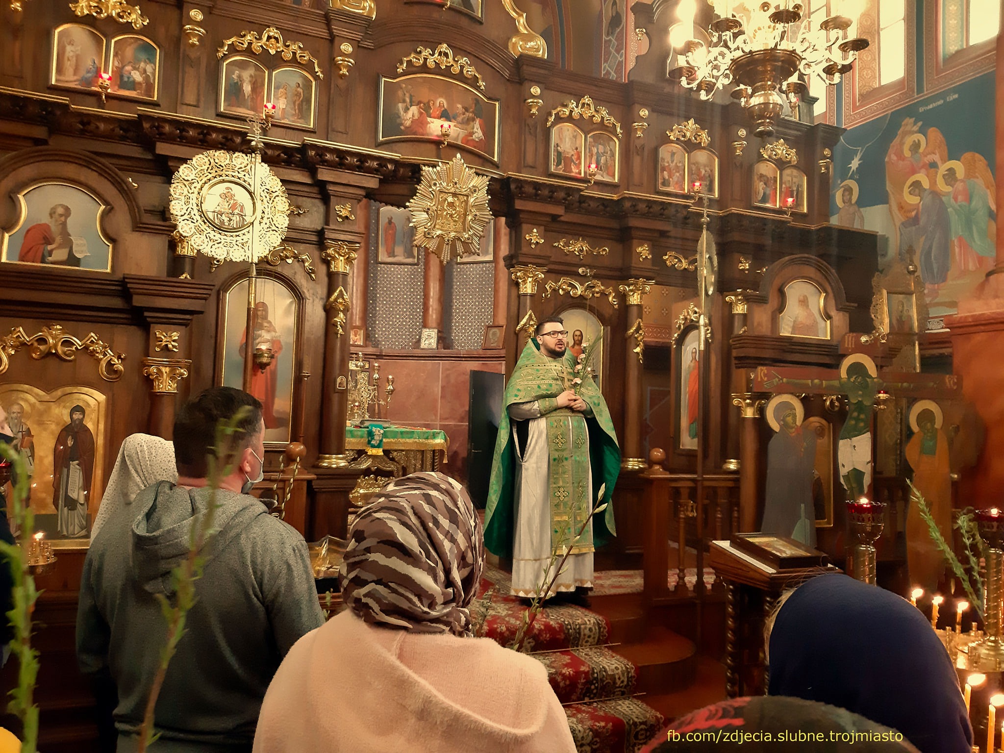 Wnętrze bogato zdobionej cerkwi z wiernymi i duchownym w szatach liturgicznych podczas nabożeństwa.