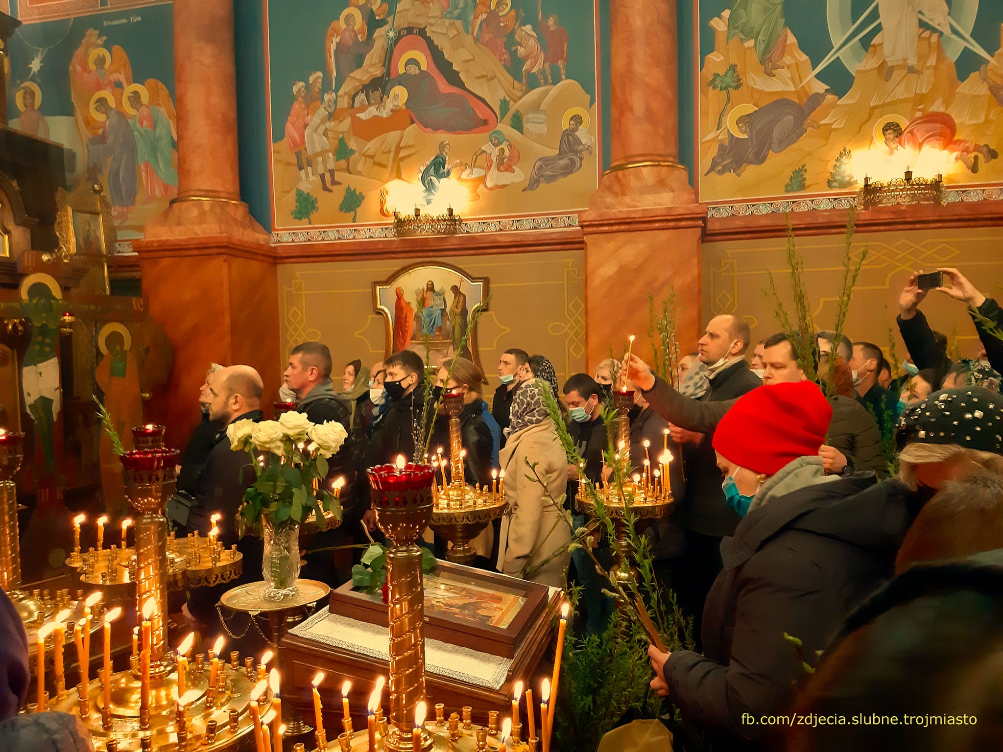 Wnętrze cerkwi podczas uroczystości religijnej: wierni trzymają zapalone świece i gałązki, w tle malowidła na ścianach i kolumnach.