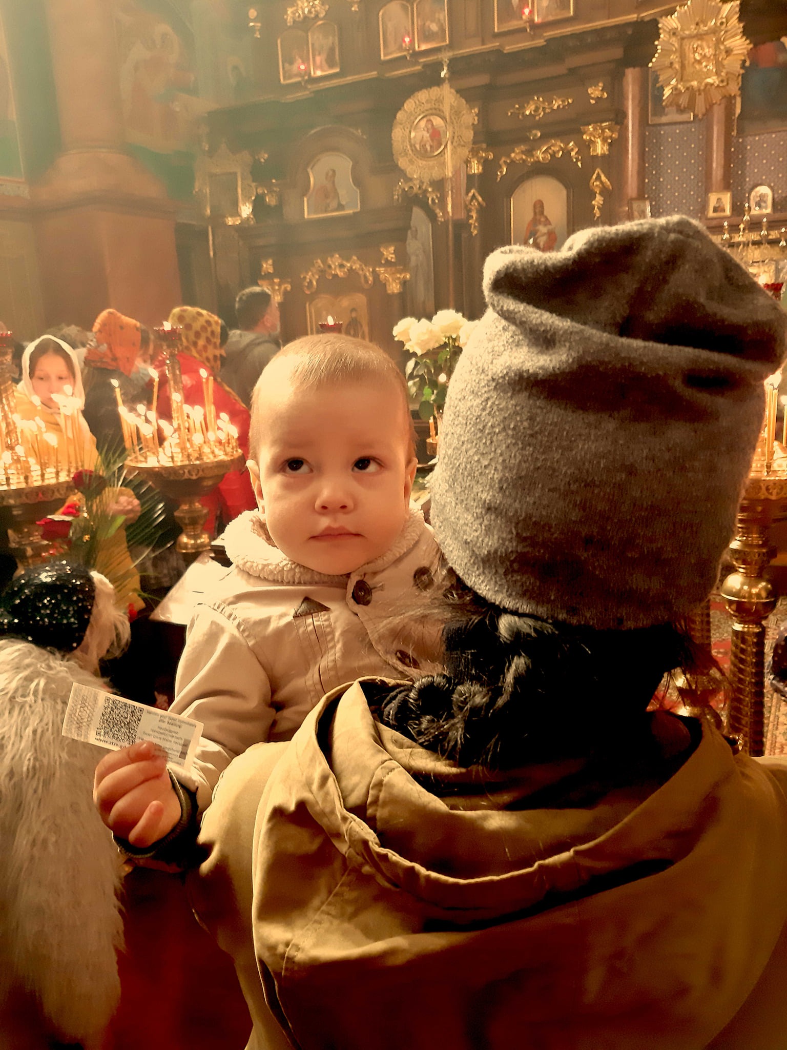 Dziecko na ramionach dorosłego w cerkwi, trzymające bilet, w tle ikony i świece.