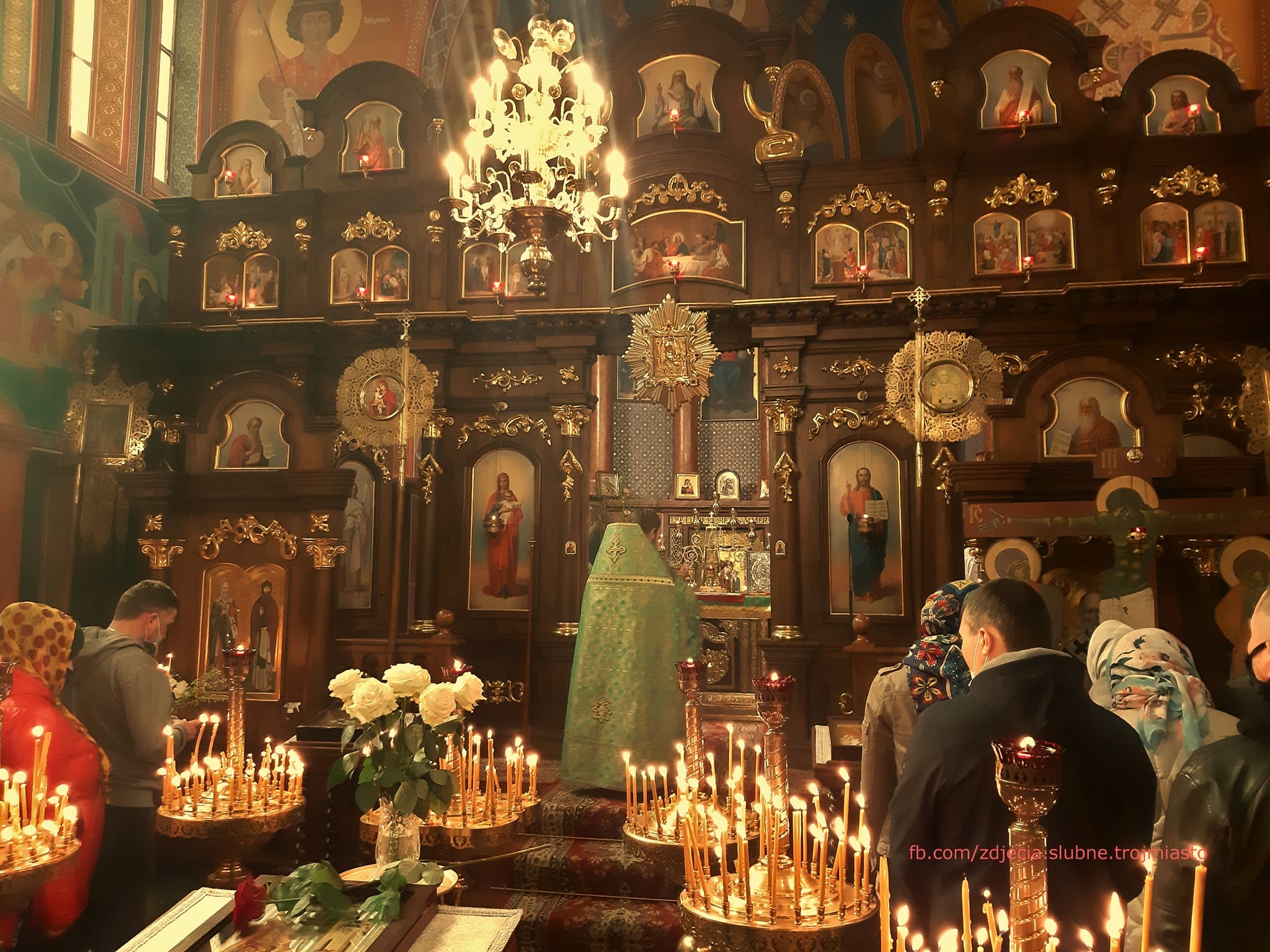 Wnętrze cerkwi z ikonami, świecami i wiernymi podczas nabożeństwa, widoczny duchowny w zielonej szacie przed ikonostasem.