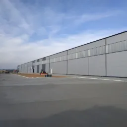 Rozległa, szara powierzchnia betonowa przed budynkiem przemysłowym z widocznymi maszynami budowlanymi w tle, pod błękitnym, zachmurzonym niebem.