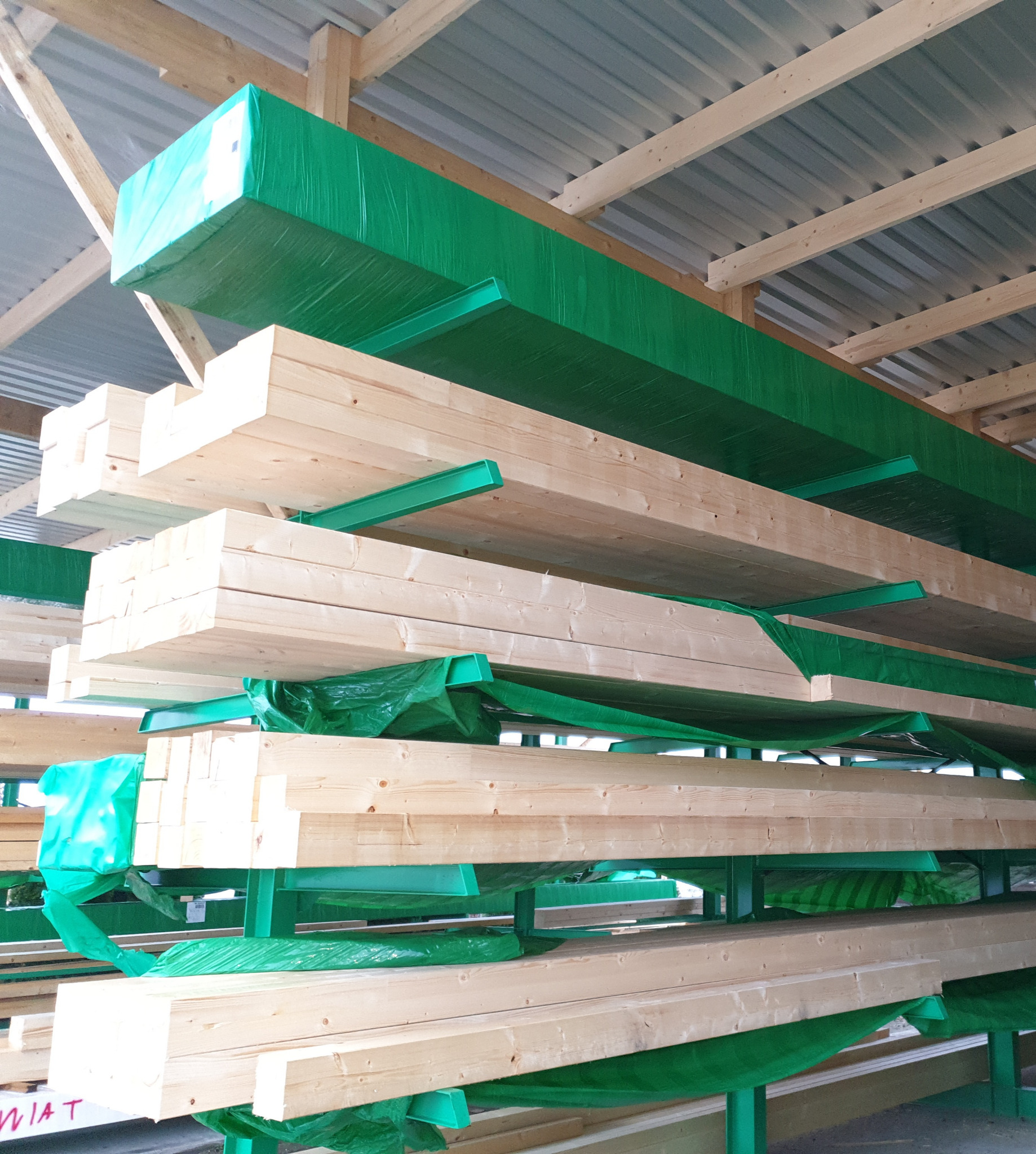 Stosy kantówki z jasnego drewna, częściowo owinięte zieloną folią ochronną, ułożone na zielonych metalowych regałach w magazynie z metalowym dachem.
