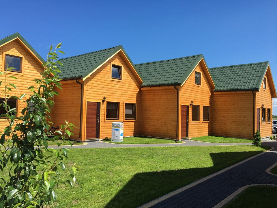 Rząd identycznych, pomarańczowych domków z drewna z zielonymi dachami, widok z poziomu trawnika, błękitne niebo w tle.