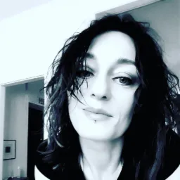 Portret kobiety z ciemnymi, potarganymi włosami i piercingiem w wardze, z delikatnym uśmiechem, na rozmytym tle wnętrza.