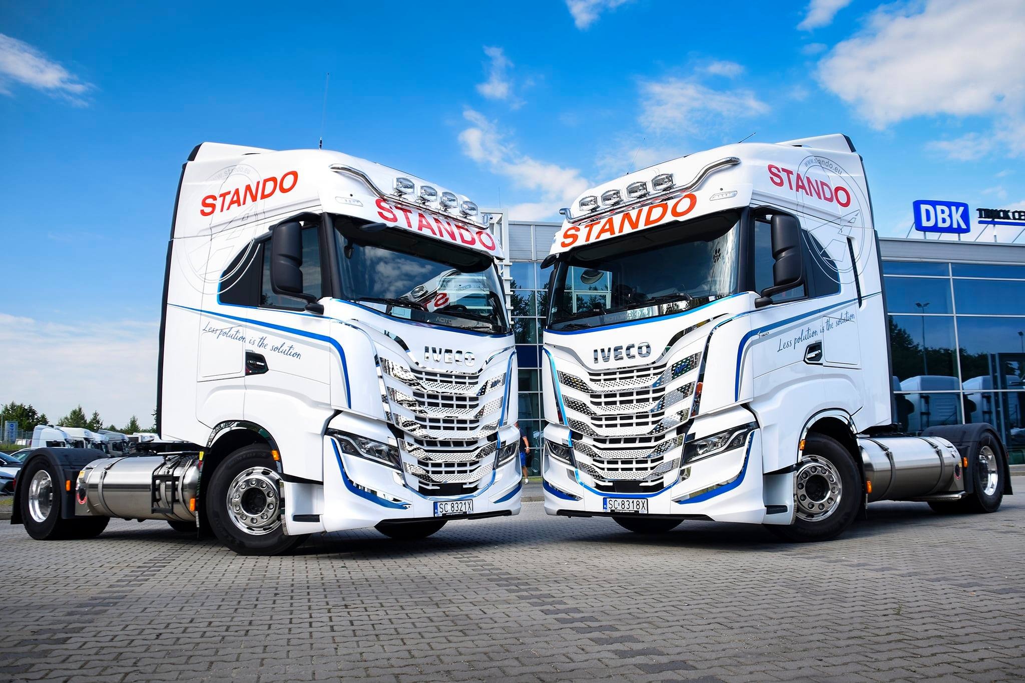 Dwie białe ciężarówki marki Iveco z logo firmy STANDO ustawione przodem do siebie na tle błękitnego nieba i nowoczesnego budynku z przeszkloną fasadą.