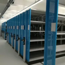 Przesuwne regały magazynowe w jasnym pomieszczeniu, z niebieskimi panelami i czarnymi uchwytami do przesuwania, widoczne puste p&oacute;łki.