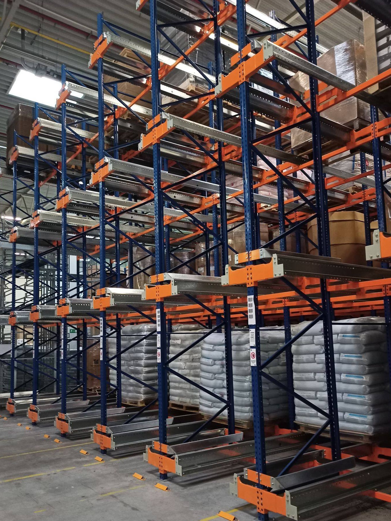 Wysokie, niebiesko-pomarańczowe regały magazynowe z paletami i workami, widok z dołu w oświetlonym magazynie.