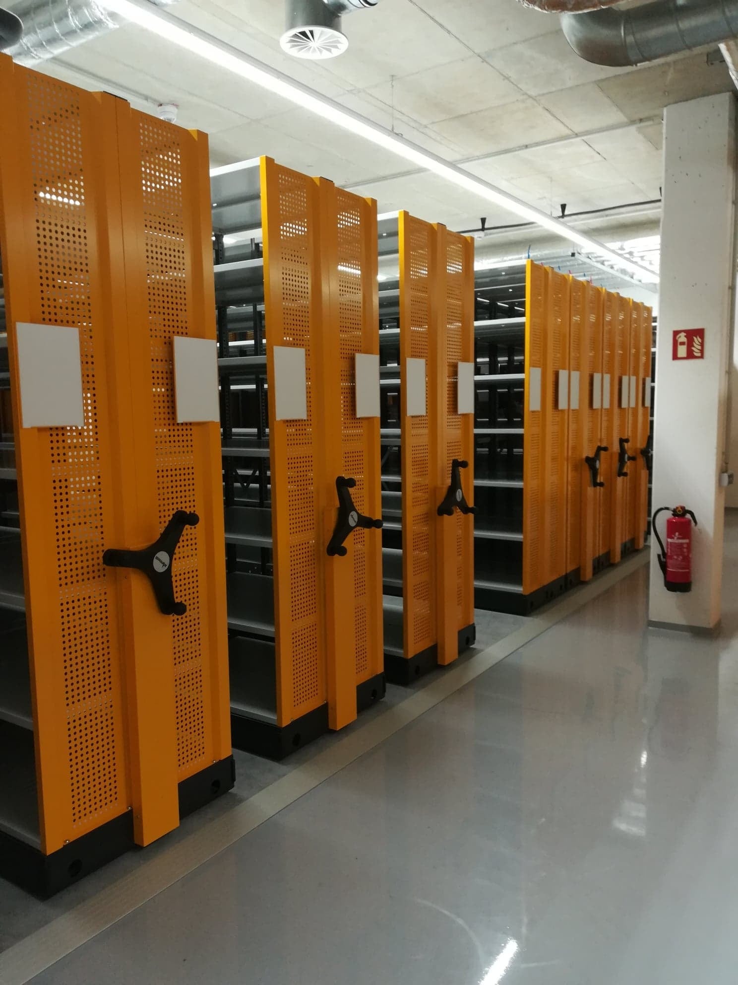 Rząd pomarańczowych, perforowanych regałów przesuwnych w magazynie z betonowym sufitem, widoczny czarny uchwyt do przesuwania, na ścianie gaśnica.