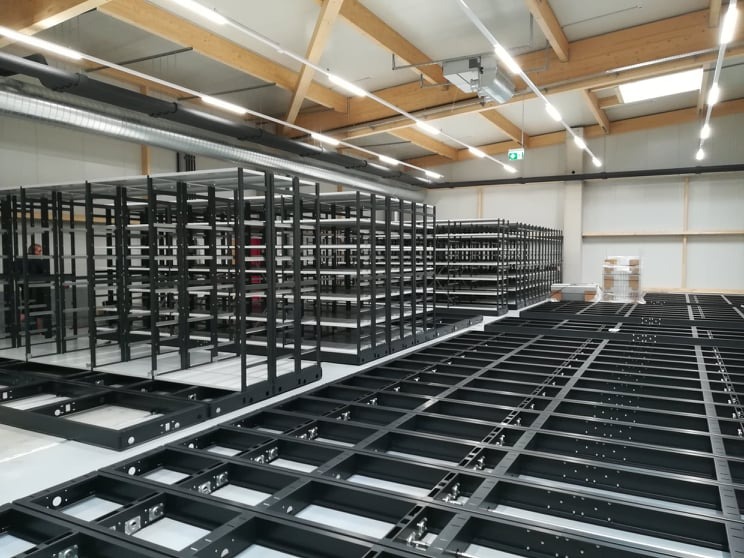 Montaż czarnych, metalowych konstrukcji regałowych w magazynie z drewnianą konstrukcją dachu. Widoczne elementy podłogowe i gotowe moduły regałów.