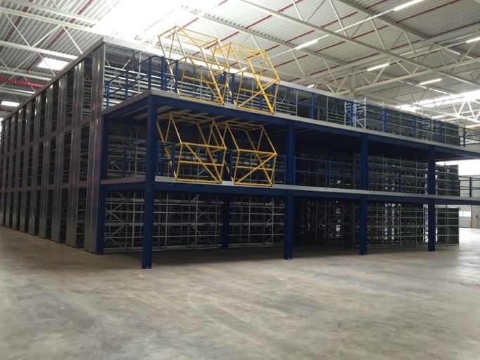 Wielopoziomowa konstrukcja magazynowa z metalowymi regałami pomalowanymi na niebiesko i szaro, z żółtymi elementami zabezpieczającymi na górnych poziomach, w jasnym magazynie.