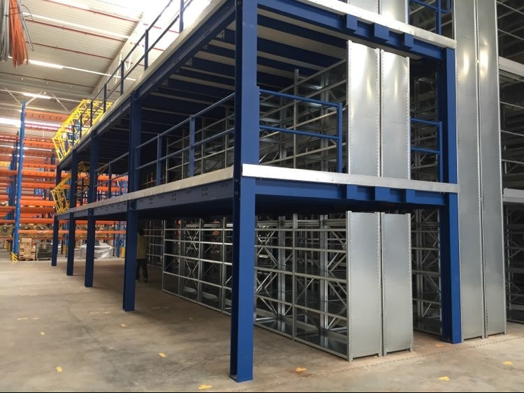 Wielopoziomowy system regałów magazynowych z niebieską konstrukcją nośną i metalowymi półkami, widok z dołu, oświetlenie magazynowe w tle.