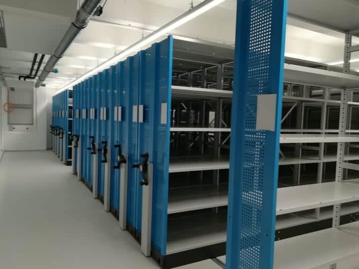 Przesuwne regały magazynowe w jasnym pomieszczeniu, z niebieskimi panelami i czarnymi uchwytami do przesuwania, widoczne puste półki.