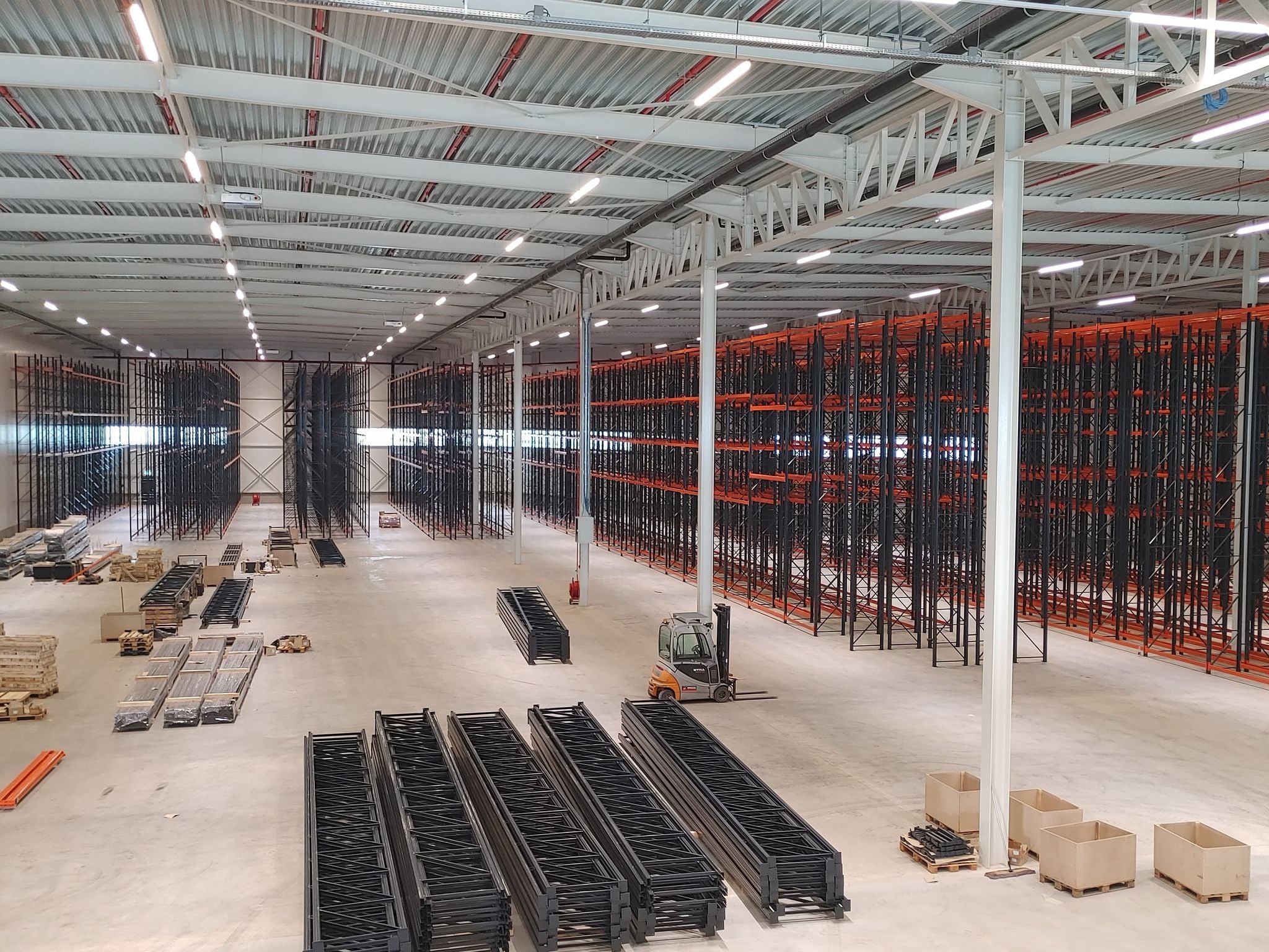 Montaż regałów magazynowych w nowej hali, widoczne elementy konstrukcyjne, wózek widłowy STILL i oświetlenie LED pod sufitem.