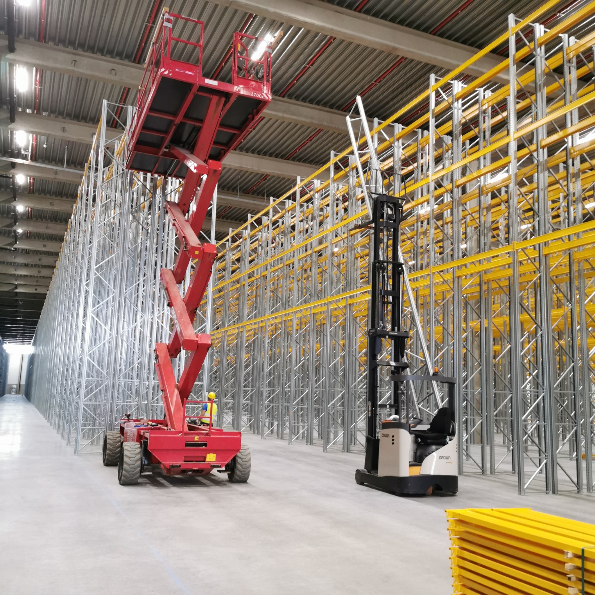 Montaż regałów magazynowych z użyciem podnośnika nożycowego i wózka widłowego w hali magazynowej. Widoczne metalowe ramy i żółte poprzeczki.