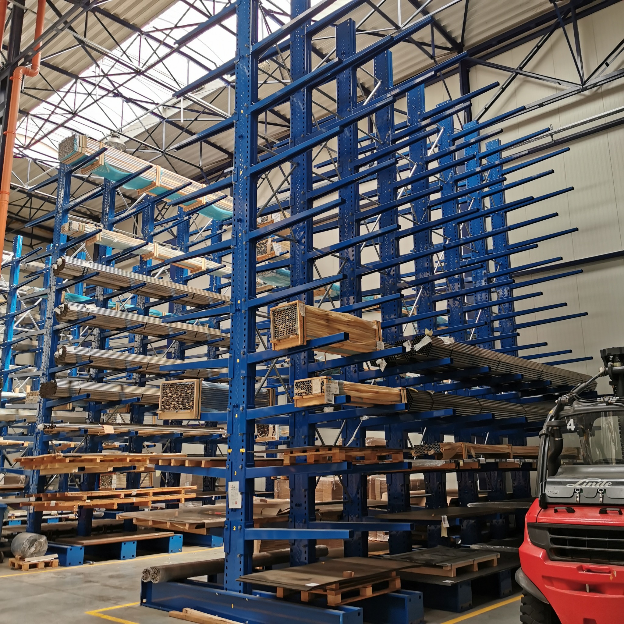 Niebieski regał magazynowy z ramionami, wypełniony różnymi materiałami, w tle widoczny wózek widłowy Linde.