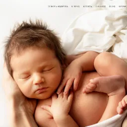 Śpiący noworodek w delikatnych dłoniach, otulony miękką białą tkaniną, z widocznym logo fotografa w lewym górnym rogu i menu strony internetowej u góry.