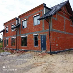 Budynek w trakcie budowy z czerwonej cegły, widoczne okna z roletami zewnętrznymi, drzwi wejściowe i elementy dachu, teren wok&oacute;ł budynku częściowo uprzątnięty, z pozostałościami materiał&oacute;w...