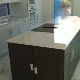 Nowoczesna wyspa kuchenna z czarnymi frontami i białym blatem, w tle zabudowa kuchenna z białymi szafkami i sprzętem AGD, marmurowa podłoga.