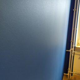 RUTAS sp. z o.o. - Świeżo pomalowana, jednolita, błękitna ściana z widocznymi rurami grzewczymi przy krawędzi i fragmentem drewnianej podłogi.