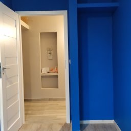 RUTAS sp. z o.o. - Pomalowany na intensywny niebieski pokój z otwartymi białymi drzwiami i panelami podłogowymi w odcieniach szarości, widoczna wnęka z półką w sąsiednim pomieszczeniu.