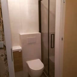 RUTAS sp. z o.o. - Nowoczesna łazienka z beżowymi płytkami, kabiną prysznicową z ciemną ramą, wiszącą toaletą i szafką z umywalką z drewnianym frontem.