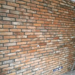 RUTAS sp. z o.o. - Ściana z cegieł klinkierowych w odcieniach czerwieni i brązu, z widocznymi fugami w kolorze grafitowym, gniazdka elektryczne przy podłodze, fragment drabiny w prawym dolnym rogu.