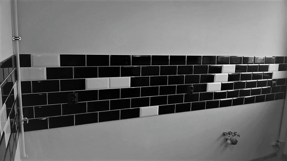 Ściana wyłożona czarnymi i białymi, błyszczącymi płytkami ceramicznymi w układzie pasowym, z widocznymi gniazdkami elektrycznymi i fragmentami instalacji wodnej.